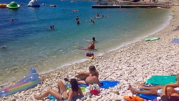 En strand med folk som badar och solar, en klippig kustlinje och ett klart, blått hav.