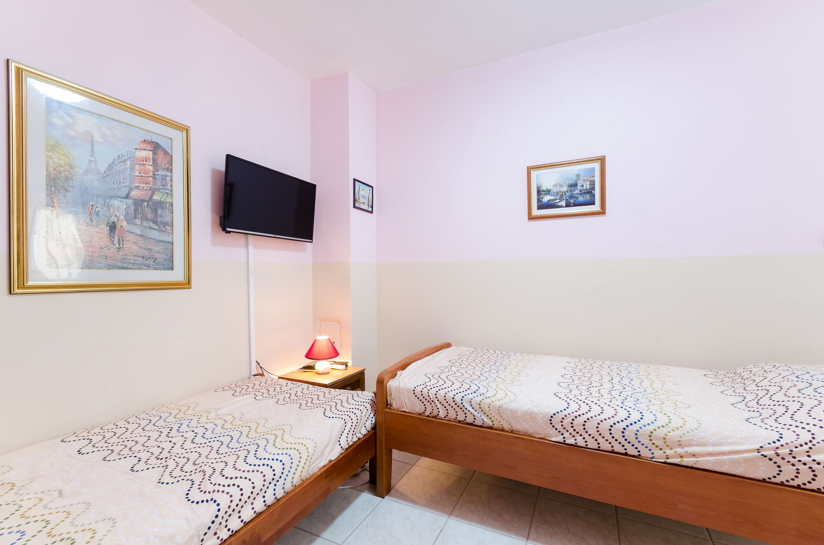 A bedroom with two single beds, a TV, a lamp, and two framed pictures on the wall.