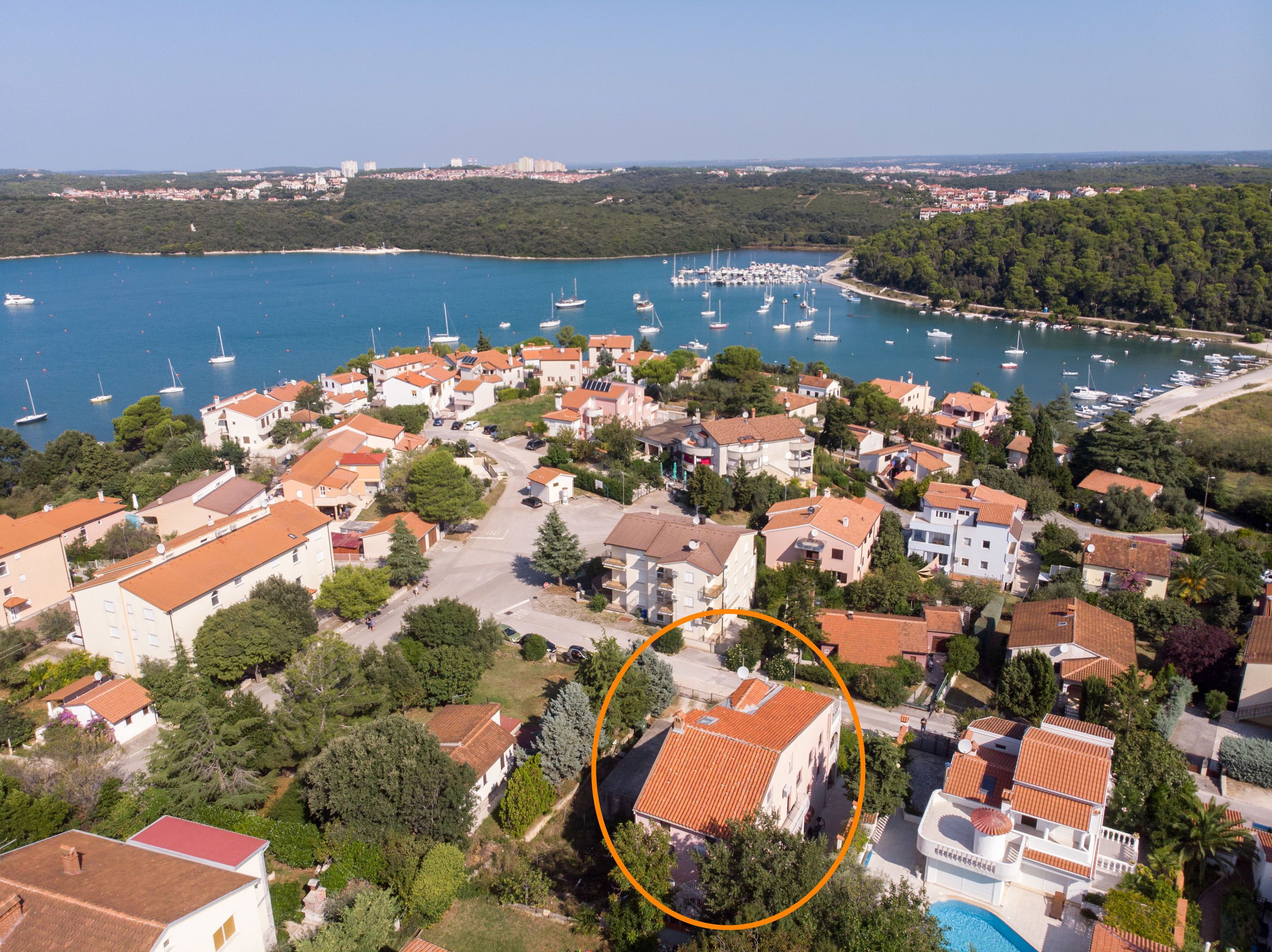 Vue aérienne d’une ville côtière dotée d’un port de plaisance, où l’on aperçoit un mélange de bâtiments résidentiels aux toits recouverts de tuiles rouges, le tout entouré de verdure et bordé par une mer bleue et limpide.