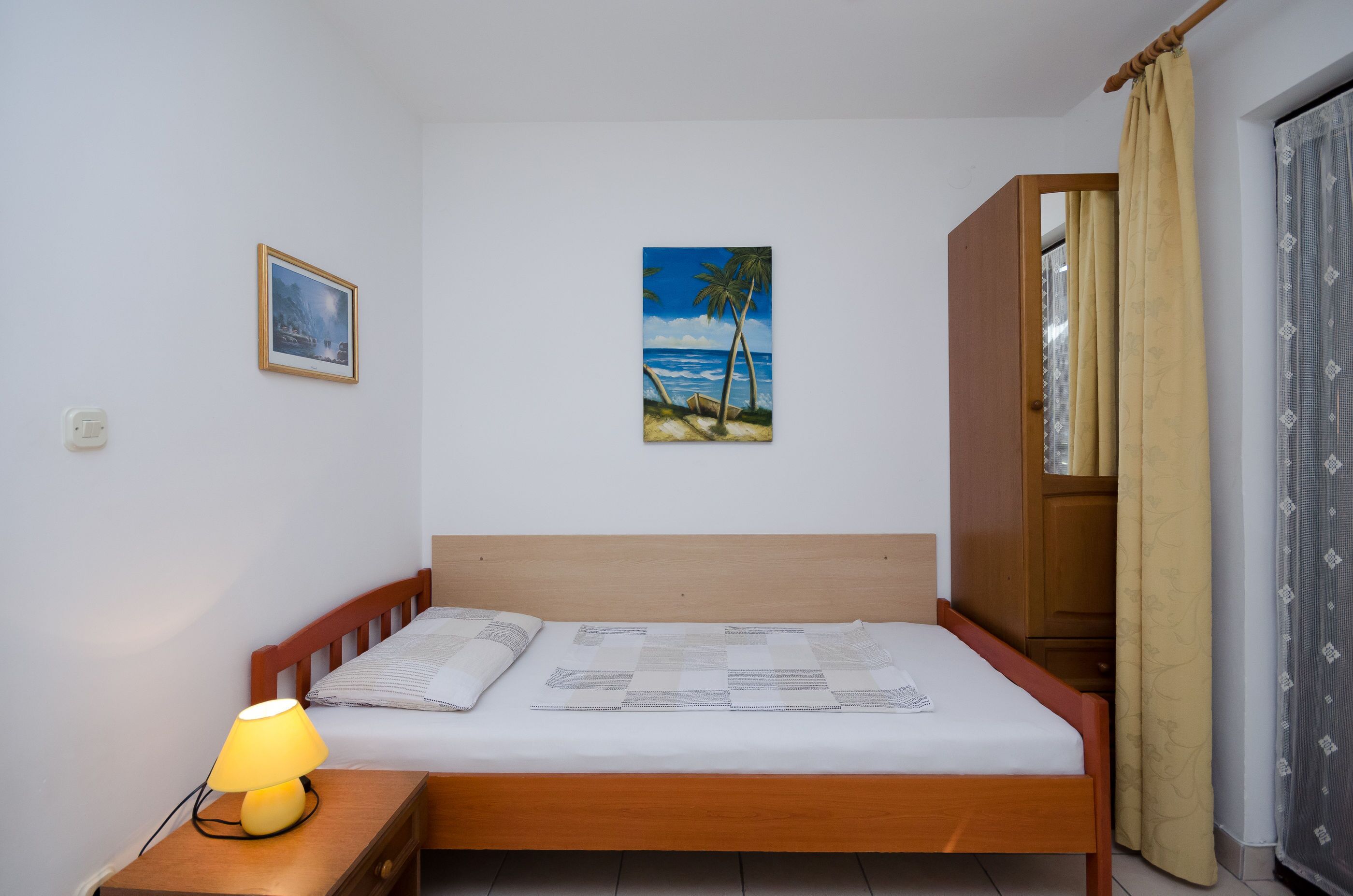 Una cama individual con cabecera de madera, una mesita de noche con lámpara, un cuadro enmarco con una escena de playa y una ventana con cortinas.