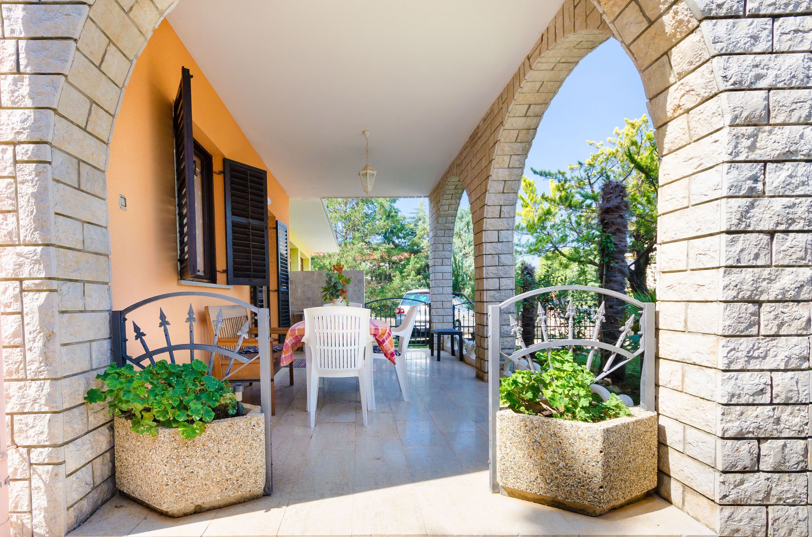 A covered outdoor area with stone arches, a table with chairs, and planters with greenery.