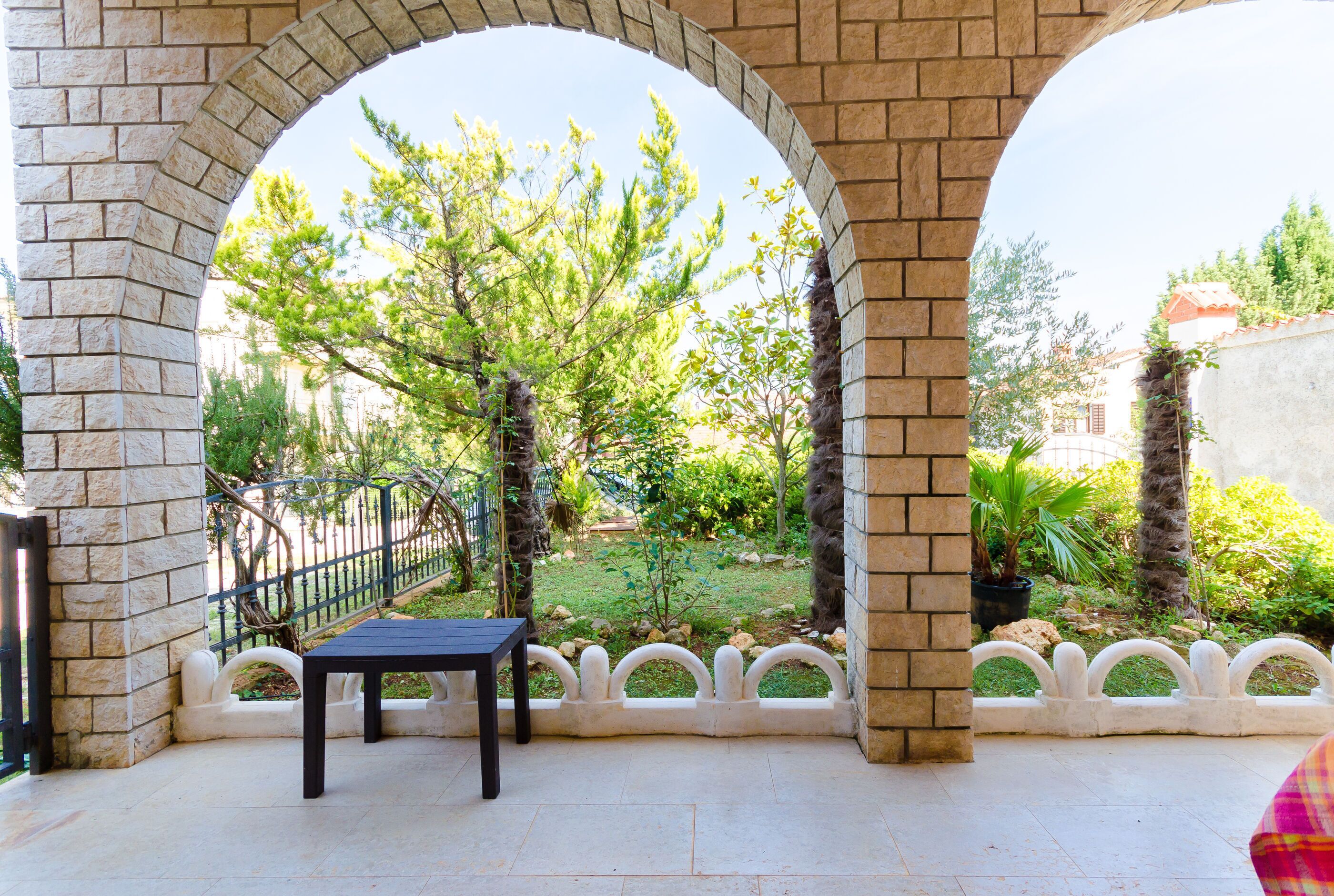 A covered patio area with a table and chairs, stone arches, and a view of a garden with trees and a fence.