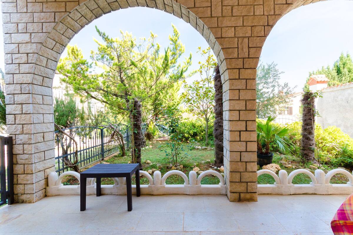 A covered patio area with a table and chairs, stone arches, and a view of a garden with trees and a fence.