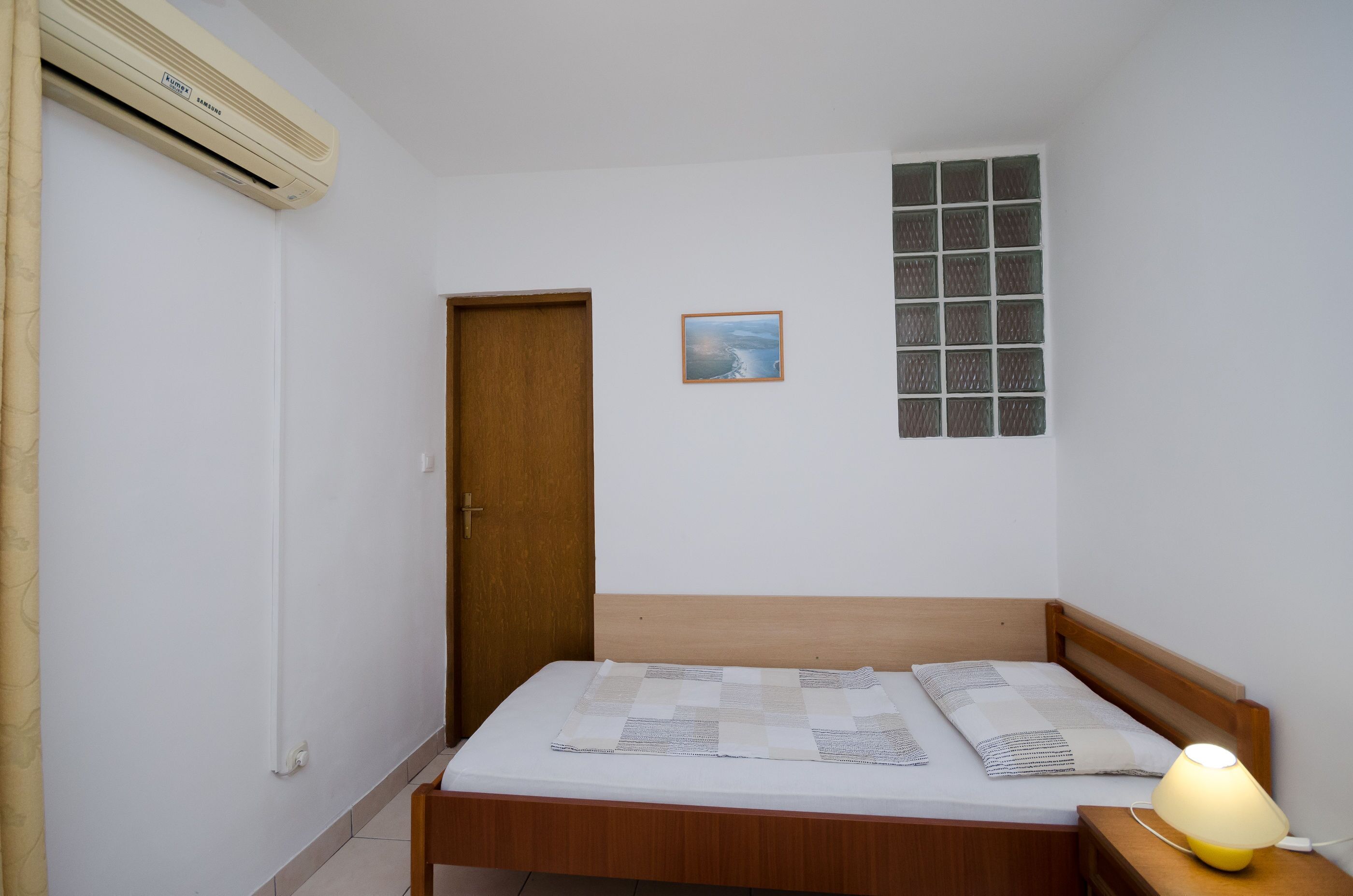 A bedroom with a wooden bed, a bedside lamp, a window with a view, and an air conditioning unit.