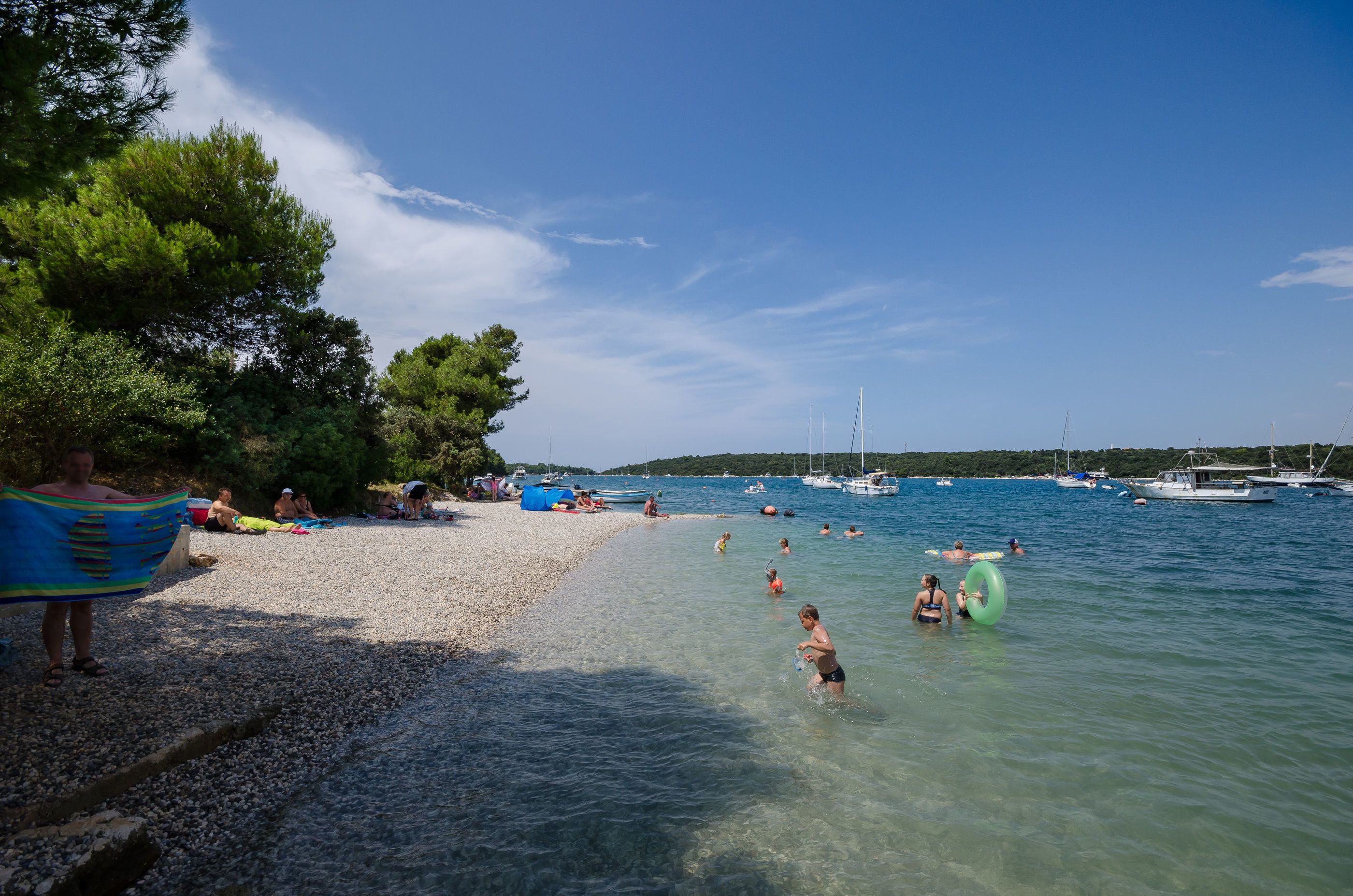 A beach with clear water, people swimming and sunbathing, and boats in the background.