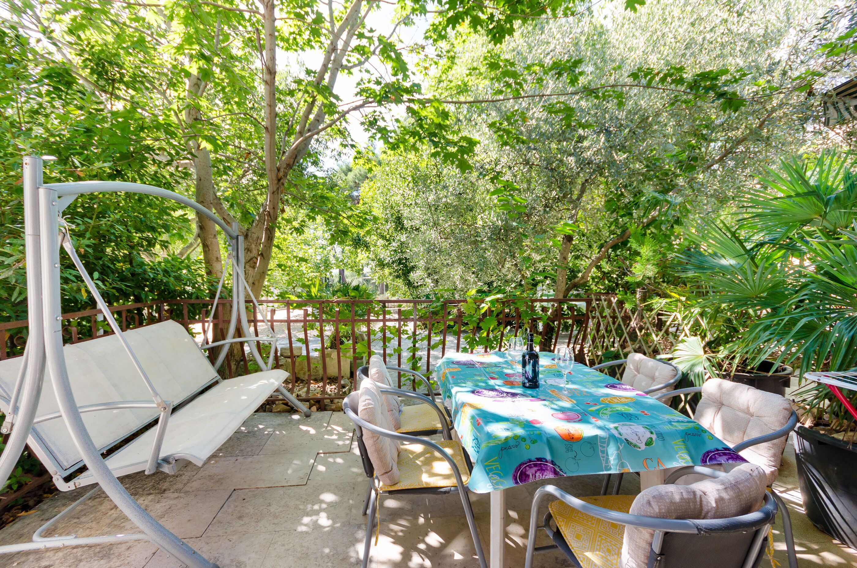 An outdoor patio with a table covered in a colorful tablecloth, surrounded by metal chairs and a swing chair.