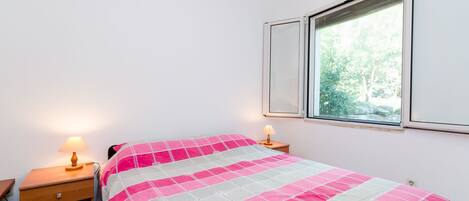 A bedroom with a bed, two bedside tables, a window, and a view of trees.