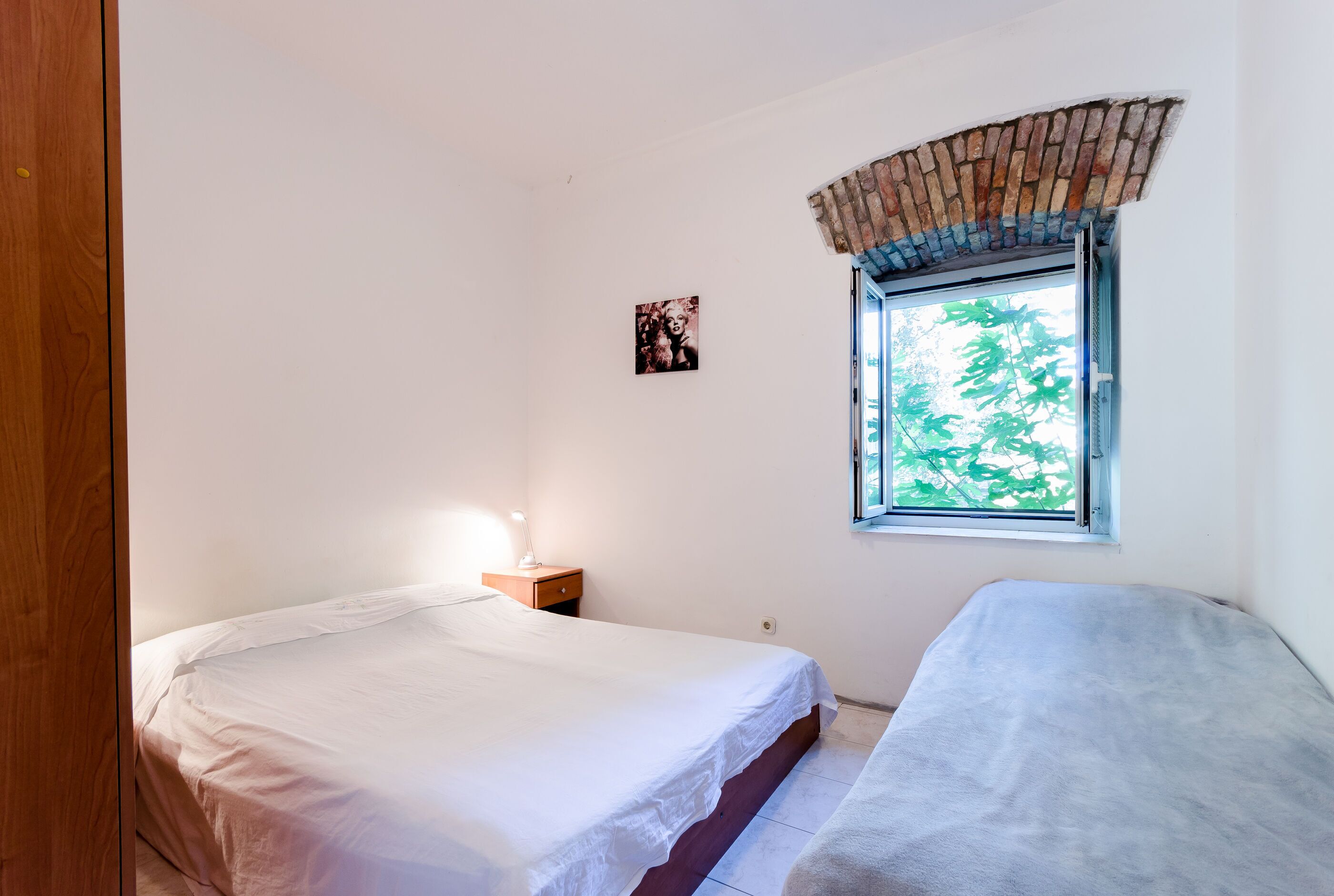 A bedroom with two beds, a window with a brick arch, a bedside table with a lamp, and a small picture on the wall.