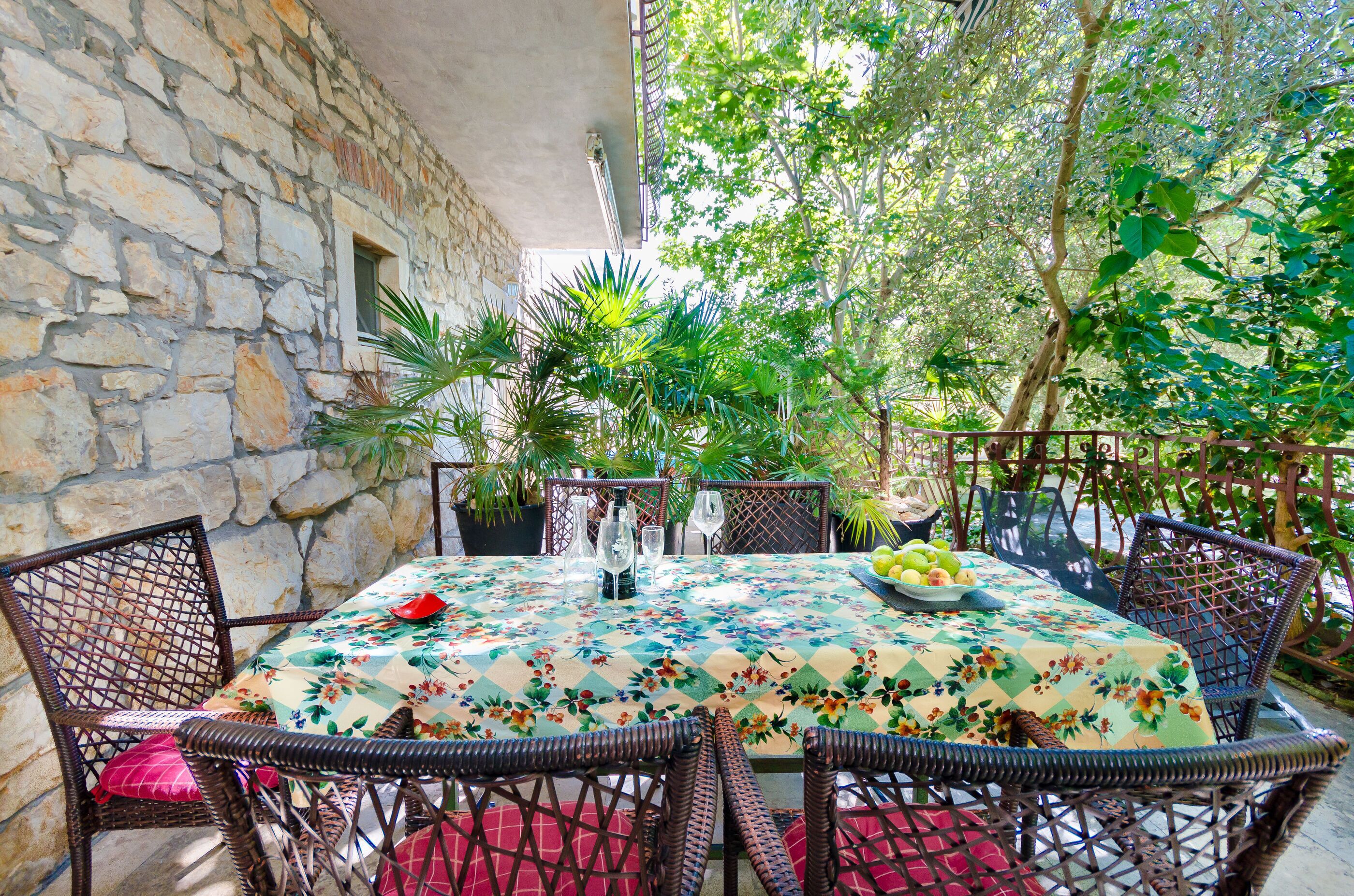 Una veranda esterna con un muro in pietra, sedie in vimini, un tavolo con tovaglia floreale e piante in vaso.
