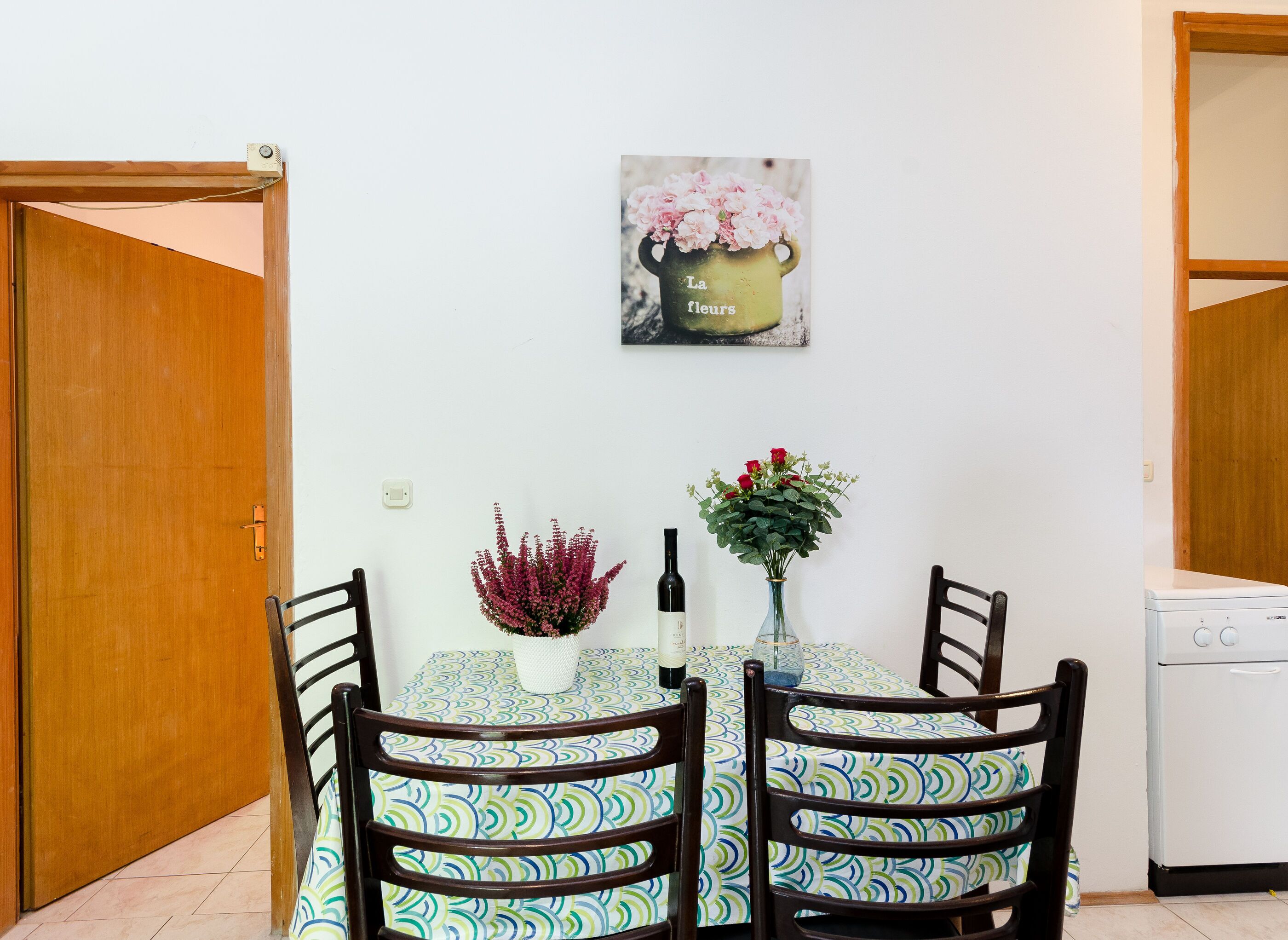 A dining area with a table set for two, surrounded by chairs with a patterned tablecloth. There is a vase with flowers on the table, and a painting above it.