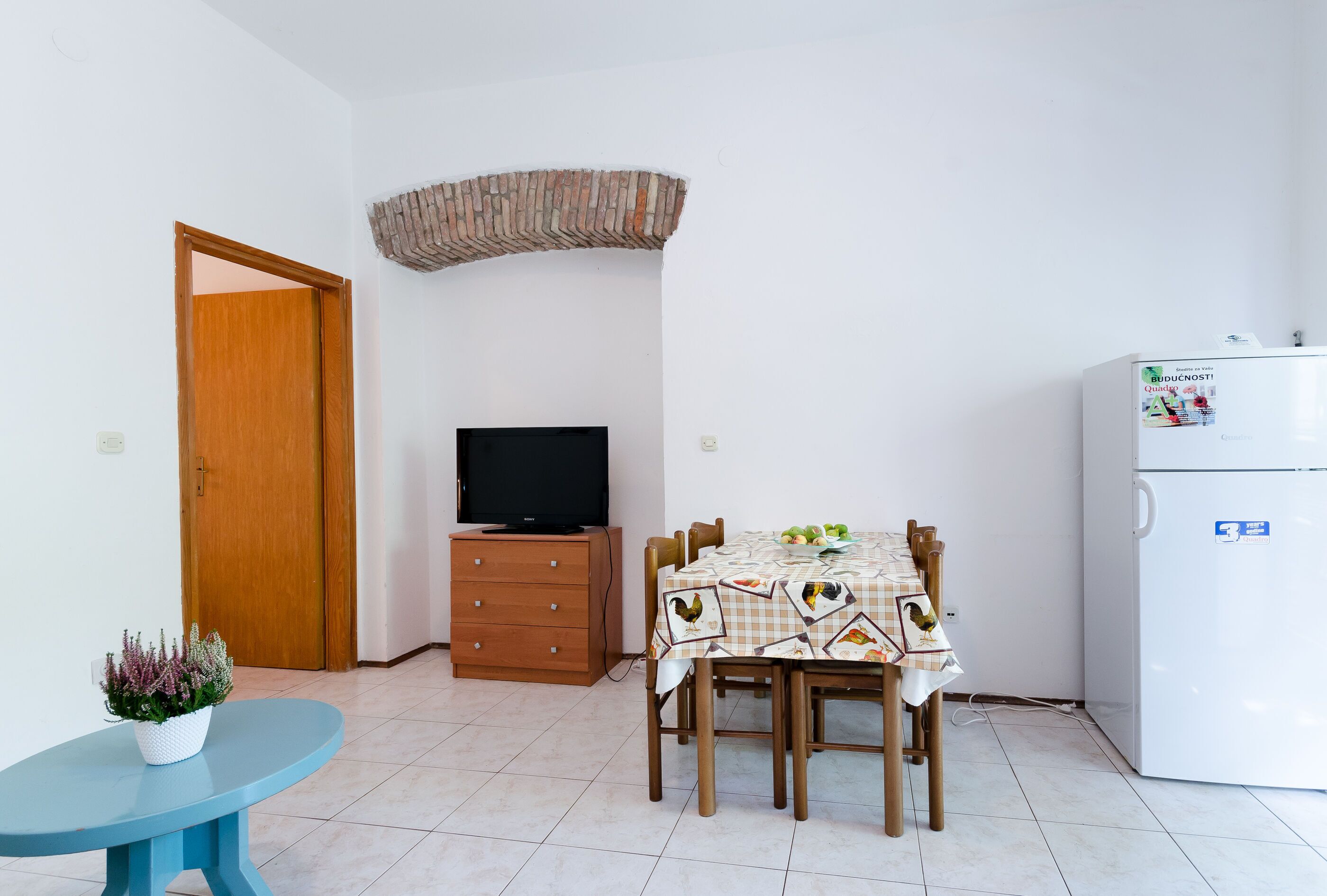 Habitació petita amb una porta de fusta, una televisió sobre un moble de fusta, una taula de menjador amb un mantell daurat i un frigorífic blanc.