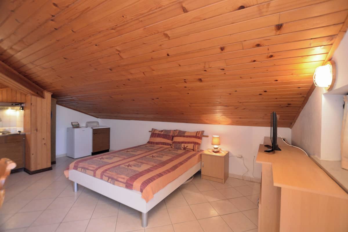 A bedroom with a wooden ceiling, a bed with a patterned blanket, a bedside table with a lamp, and a small desk with a computer.