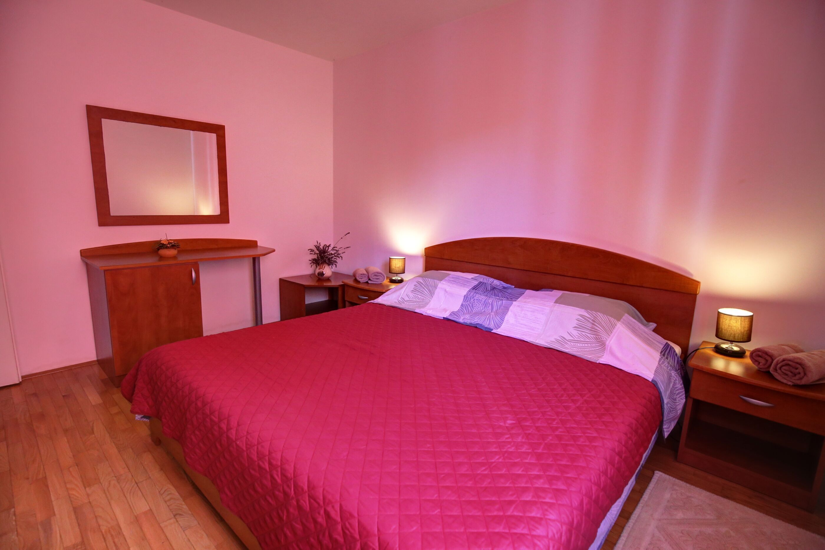 A bedroom with a large bed, a wooden nightstand, a bedside table, a mirror, and a small desk with a lamp.
