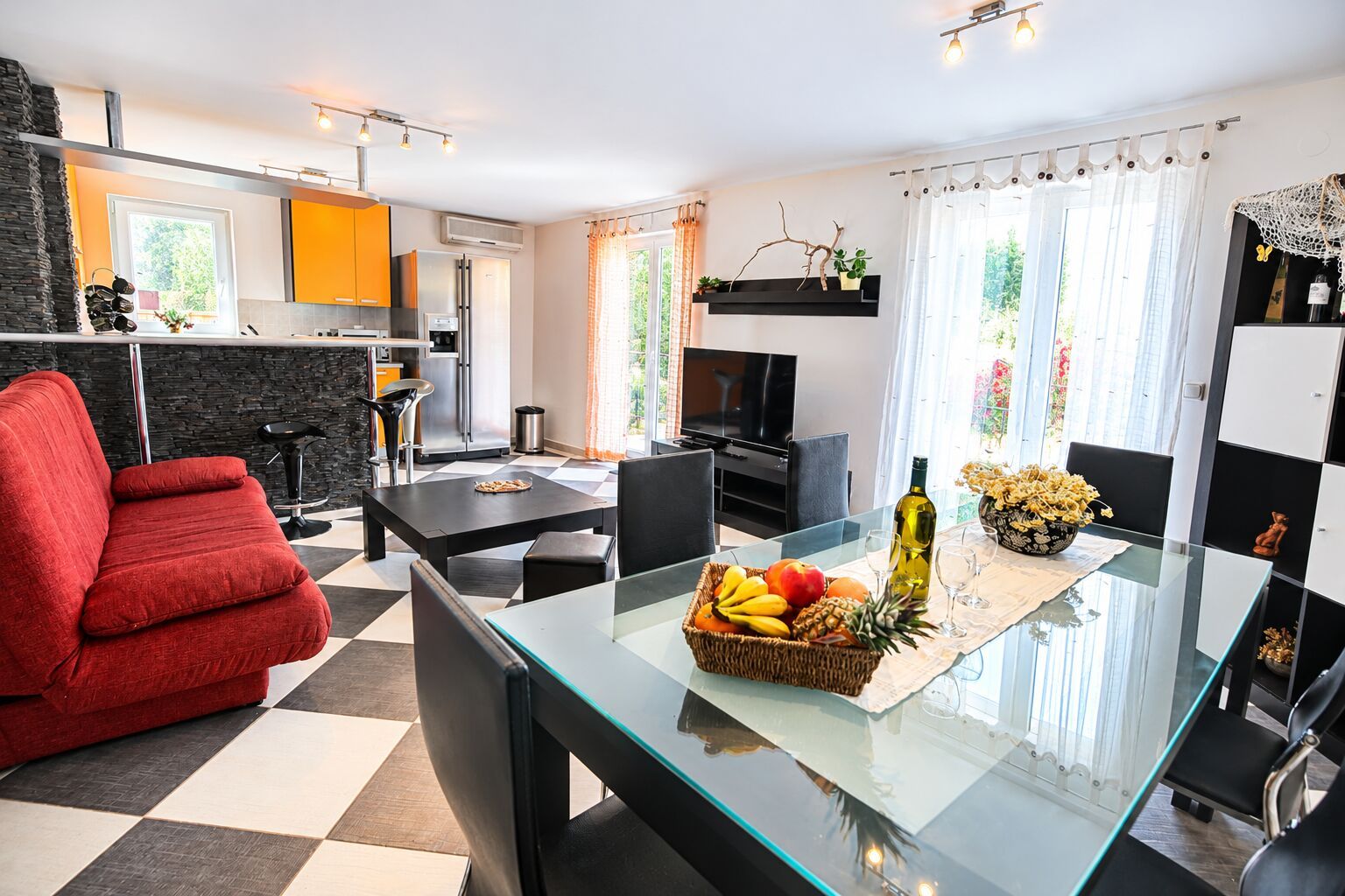 A modern living room with a glass dining table, a red sofa, and a kitchen area with a black countertop and a built-in stove.