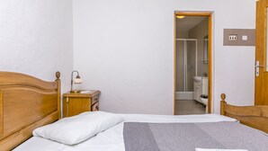 A neatly made bed with white and gray bedding, a wooden headboard, a bedside table with a lamp, and a bathroom visible through an open door. - Apartments Ivat (Rogoznica)