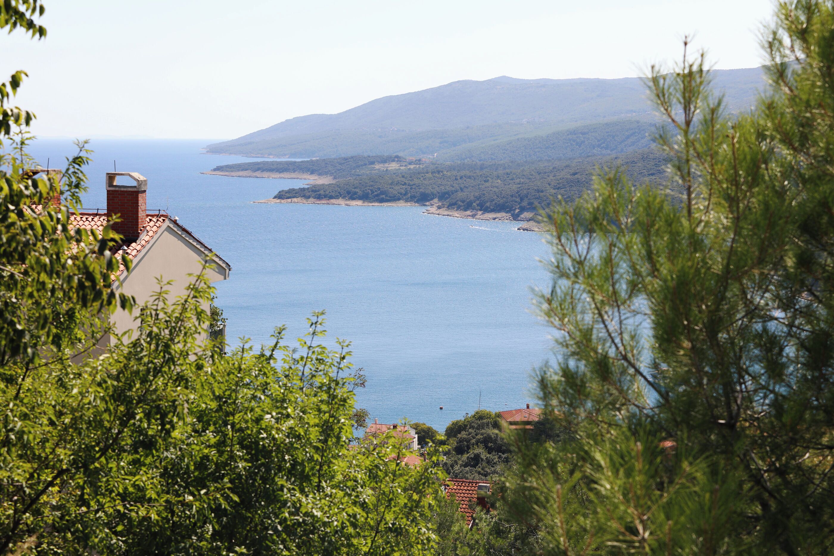 A coastal view with a building featuring a red-tiled roof, surrounded by greenery and overlooking a calm sea.