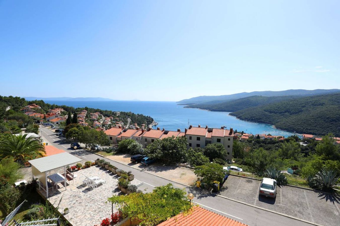 Un pueblo costero con casas escalonadas, una zona de aparcamiento pavimentada y vistas al mar y colinas.
