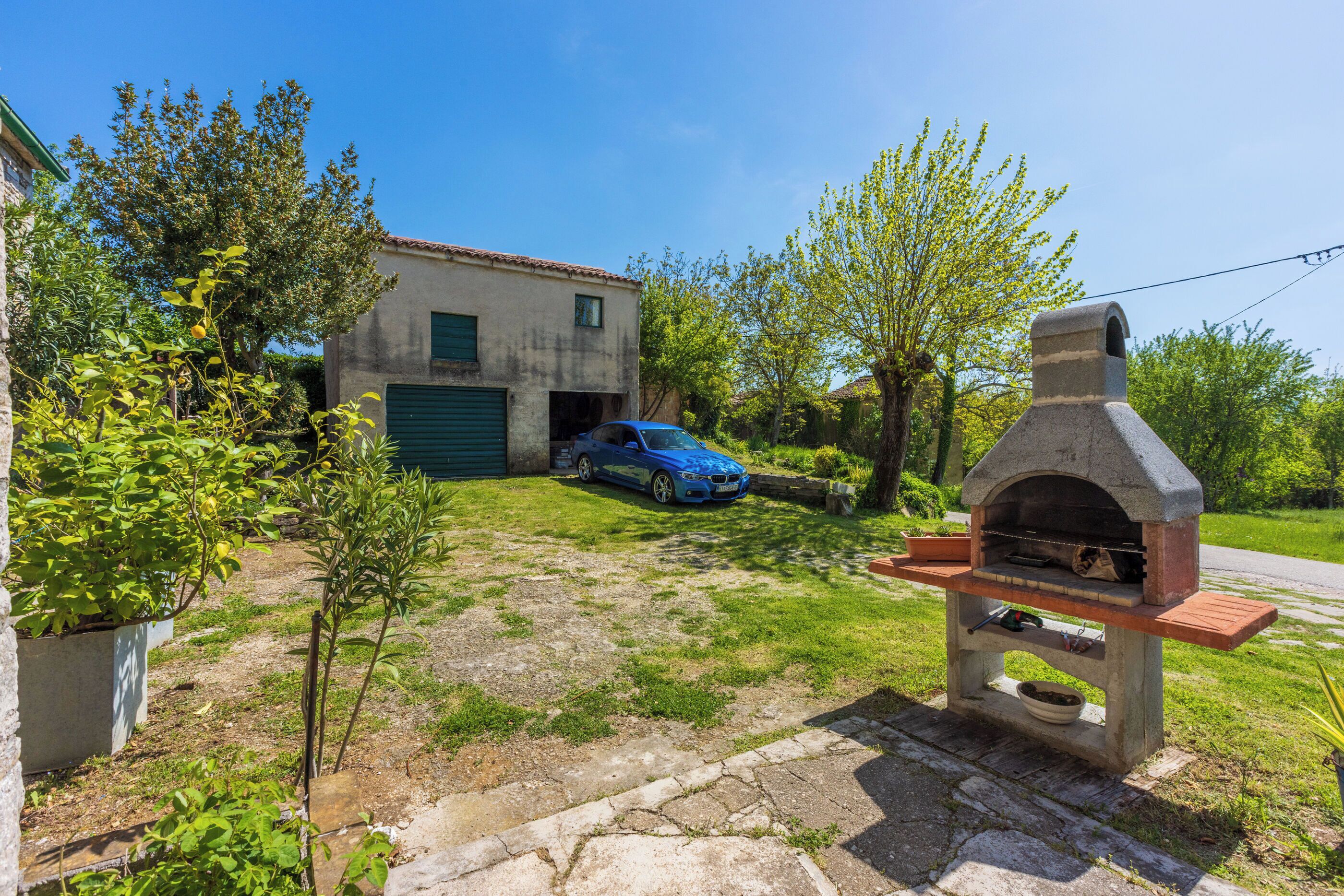 Un quartier résidentiel avec un four extérieur en pierre, une voiture bleue garée devant une maison et un petit arbre au premier plan.