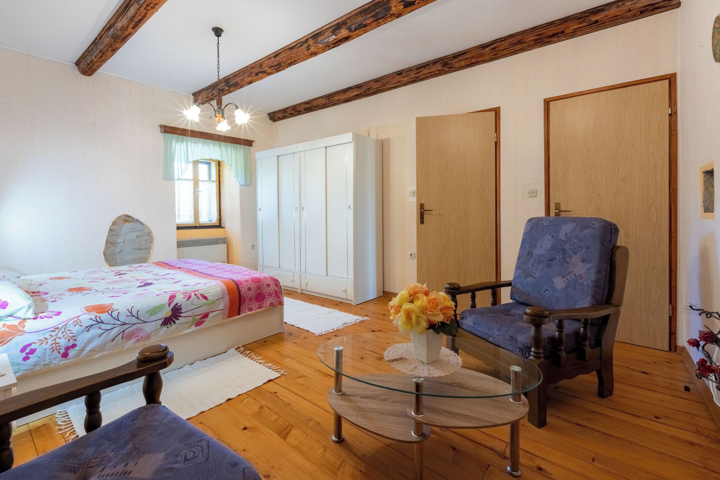 A room with a bed, a chair, a glass table with a vase of flowers, wooden floors, and a wooden ceiling beam.