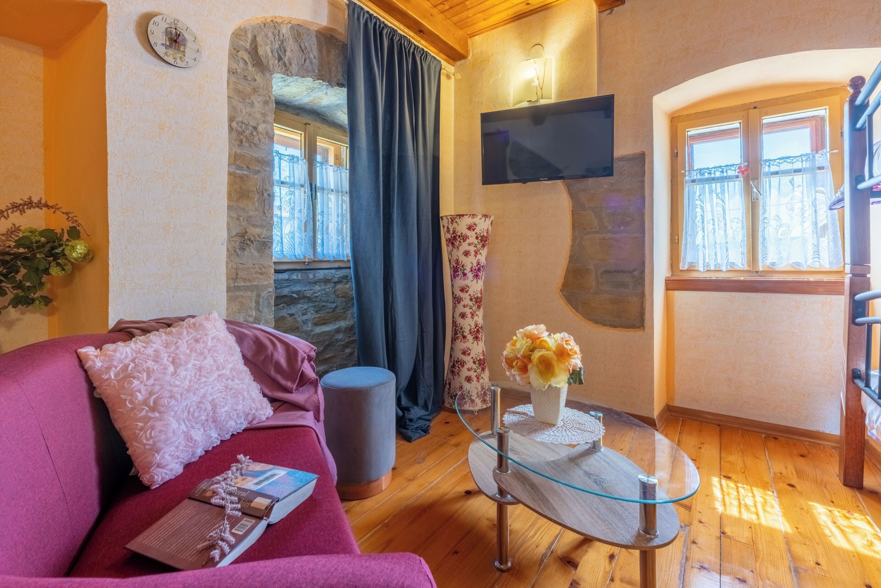 Un salon chaleureux avec un plancher en bois, un canapé rose, une table basse en verre, un mur en pierre et un téléviseur fixé au mur.