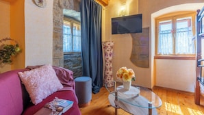 A cozy living room with a wooden floor, a pink sofa, a glass coffee table, a stone wall, and a TV mounted on the wall. - House Dorino (Lanisce Krsan)
