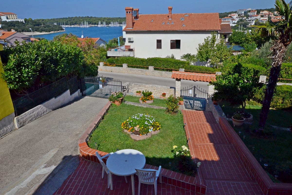 Un patio con una mesa y sillas, un pequeño jardín con flores, y vistas al mar y edificios al fondo.