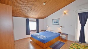 A bedroom with a wooden ceiling, a bed with a blue mattress, a nightstand, a bedside table, and a window with curtains. - Apartments and Rooms Viktorija (Pjescana uvala)