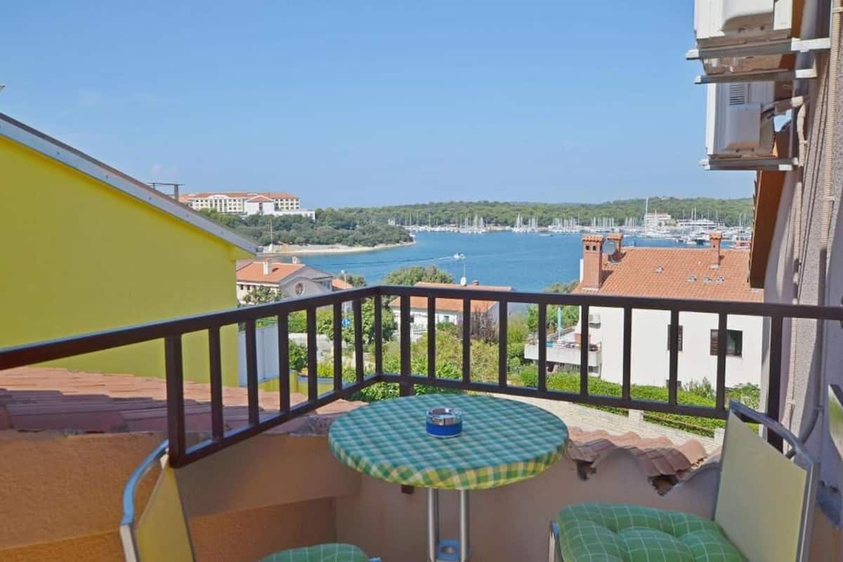 Un balcón con una mesa a cuadros y asientos acolchados verdes, con vistas a un puerto deportivo con barcos y edificios.