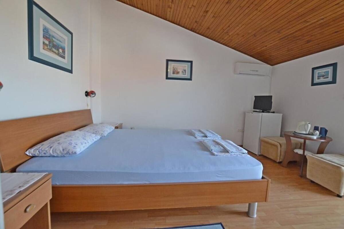 Un dormitorio con techo de madera, una cama con colchón azul, una mesita de noche, una silla, una pequeña mesa con un televisor y cuadros enmarcados en las paredes.