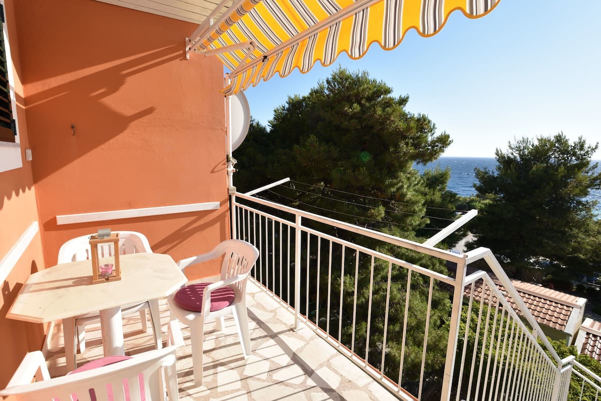 A balcony with a table and chairs, a yellow striped awning, and a view of the sea and trees.