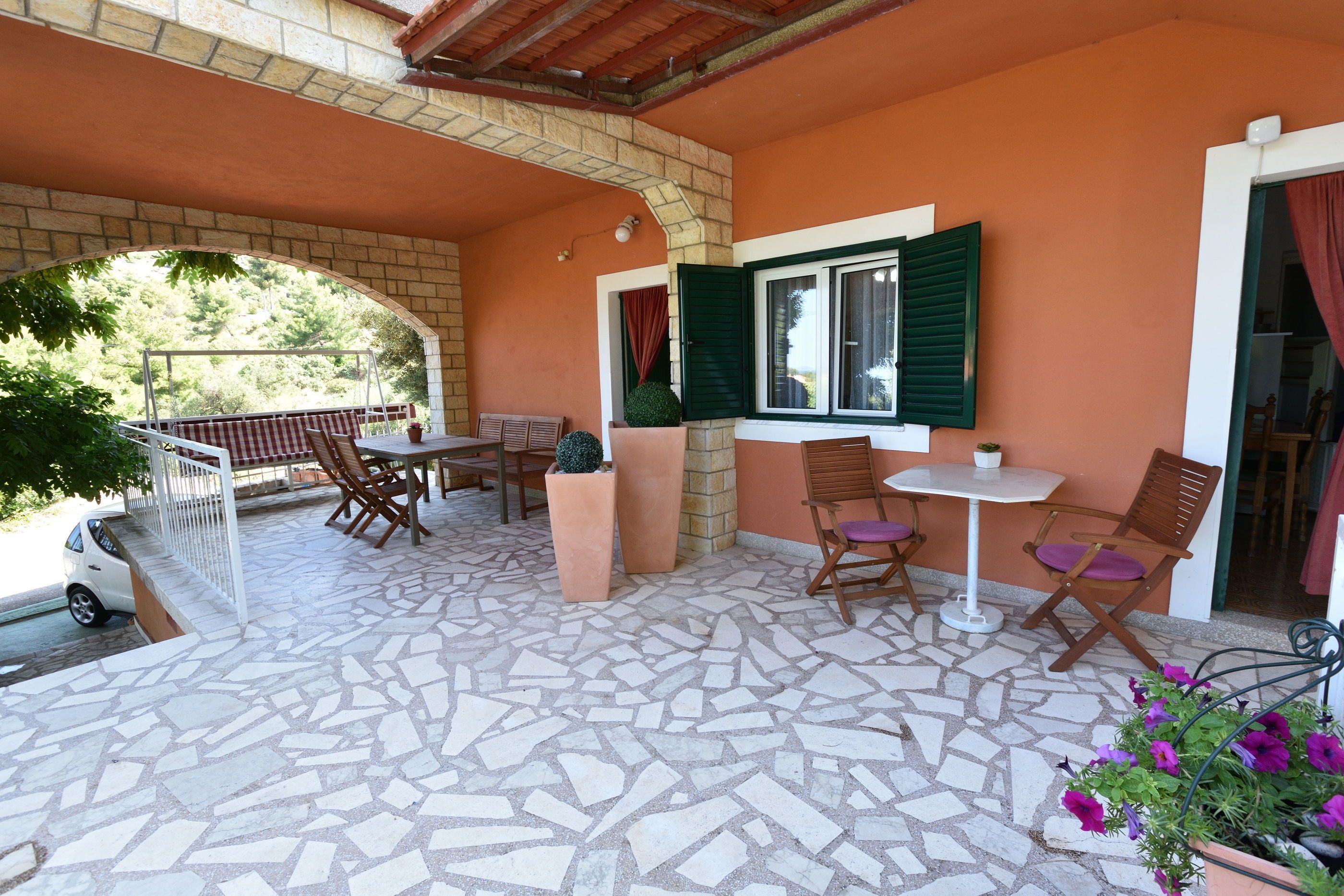 A covered patio with a mosaic floor, a table and chairs, a window with green shutters, and potted plants.