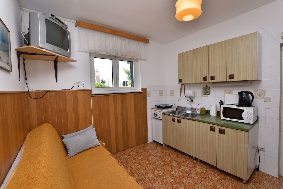 A compact kitchen with a microwave, toaster, and sink. There's a wooden cabinet and a small window with curtains.