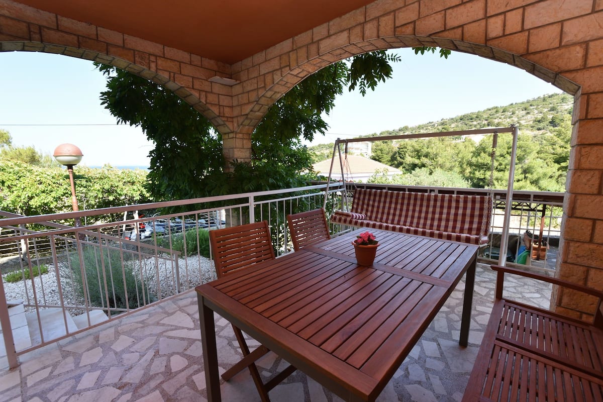 A covered outdoor patio with a wooden table and chairs, a view of a body of water, and a mountainous landscape in the background.
