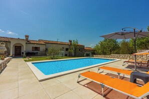 Una piscina con sillones de descanso y sombrillas, rodeada por un muro de piedra y un edificio con tejado de tejas.