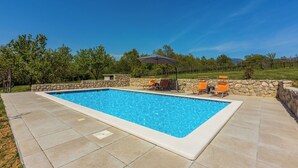 A rectangular swimming pool with a stone border, surrounded by a paved patio. There are two lounge chairs and a table with an umbrella.