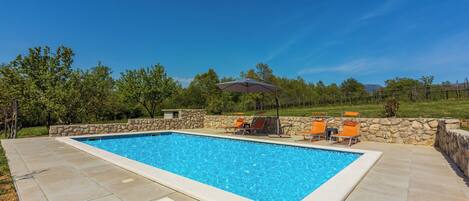 Ein rechteckiger Pool mit Steinumrandung, umgeben von einer gepflasterten Terrasse. Zwei Liegestühle und ein Tisch mit Sonnenschirm sind vorhanden.