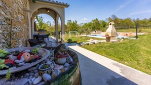 A patio area with a stone wall, a small garden with decorative items, and a view of a swimming pool and barbecue area in the background.