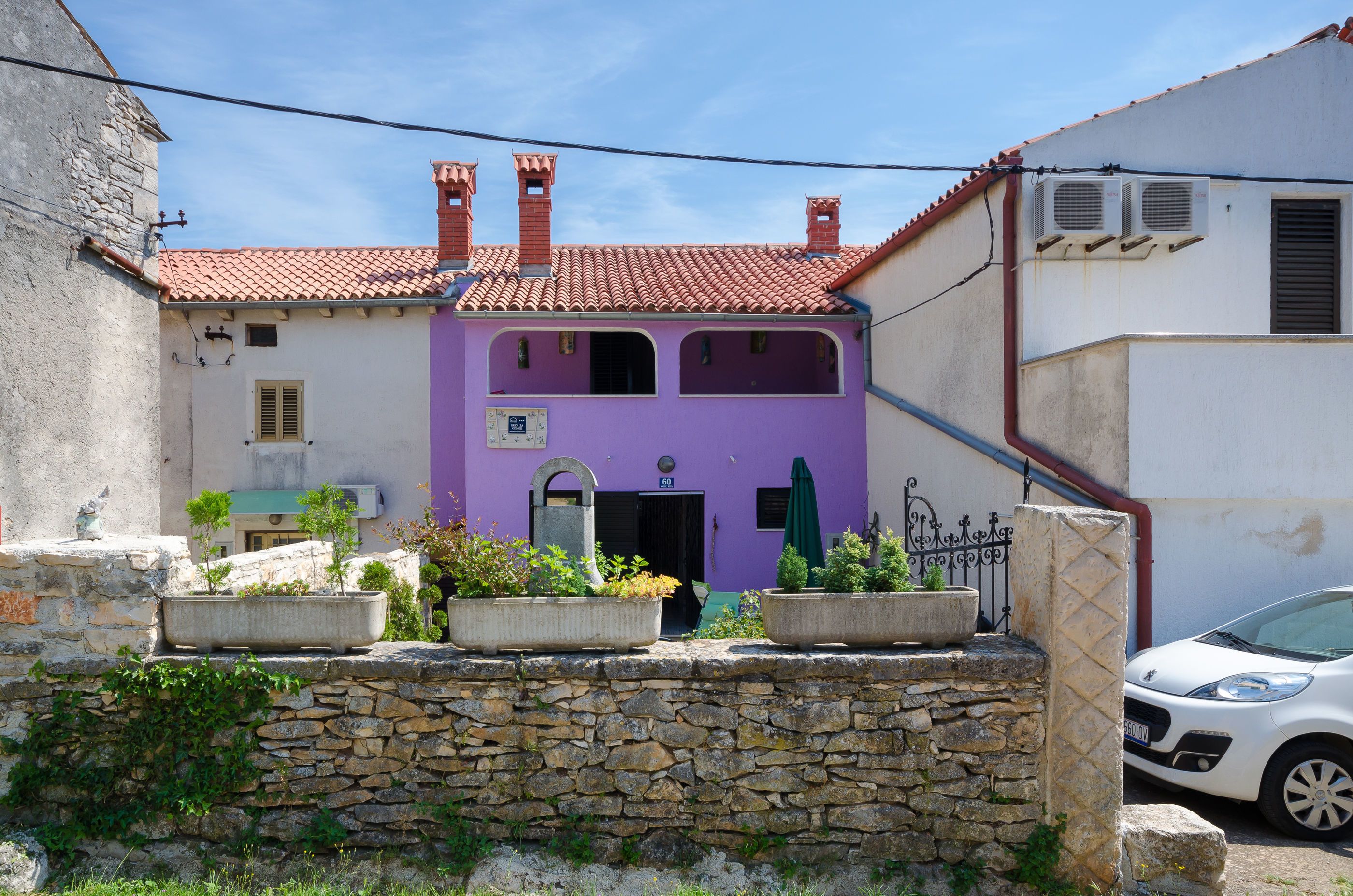 A narrow street with a purple building, stone wall, and a parked white car.