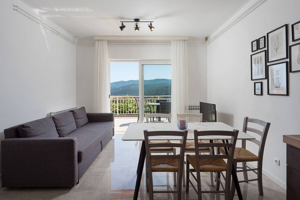 A modern living room with a sofa, dining table, chairs, and a balcony with a view of the mountains.