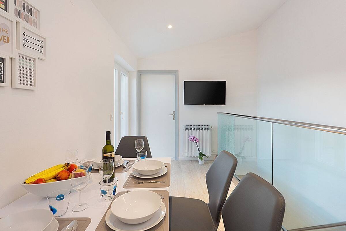 A modern dining area with a table set for two, a bottle of wine, and a bowl of fruit.