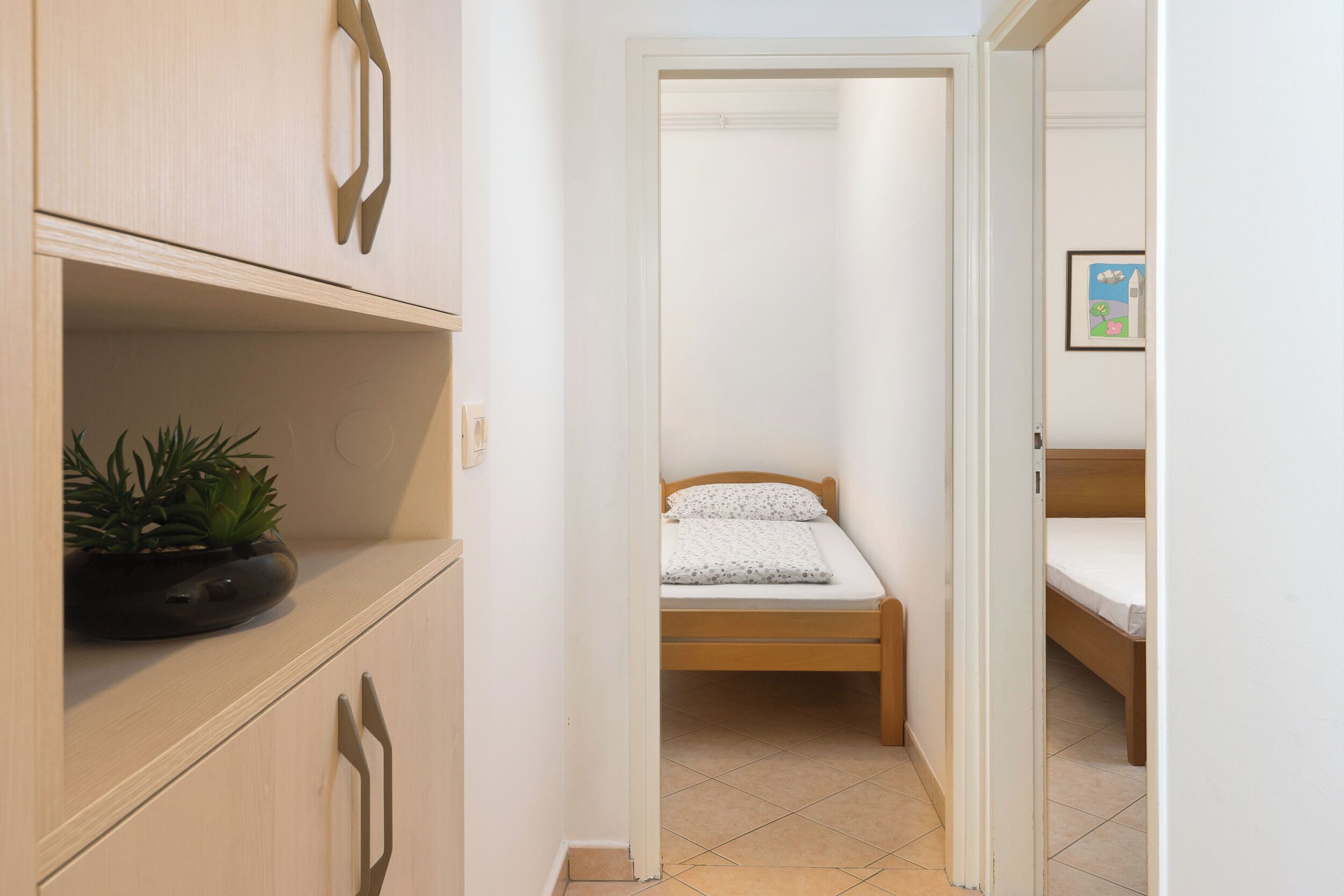 Un corridoio stretto con un armadio in legno, un letto con materasso bianco e cuscini decorati, e una piccola pianta in vaso sullo scaffale dell'armadio.