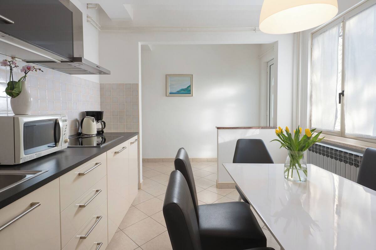 A modern kitchen with a microwave, kettle, and a vase of flowers on the counter. There is a dining table with chairs and a window with curtains.
