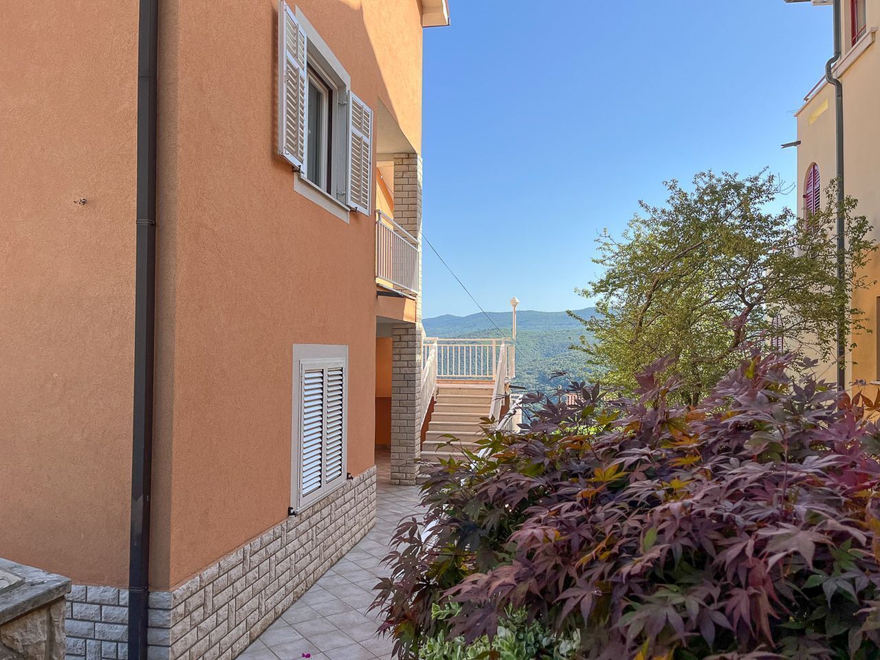 A narrow pathway leading to a building with a balcony, surrounded by vegetation and a clear view of distant mountains.
