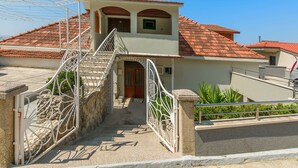 A two-story house with a red-tiled roof, a staircase leading to the entrance, and a gated driveway.
