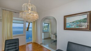 A modern living room with a wooden floor, a chandelier, a sofa, and a dining area with a view of the sea.