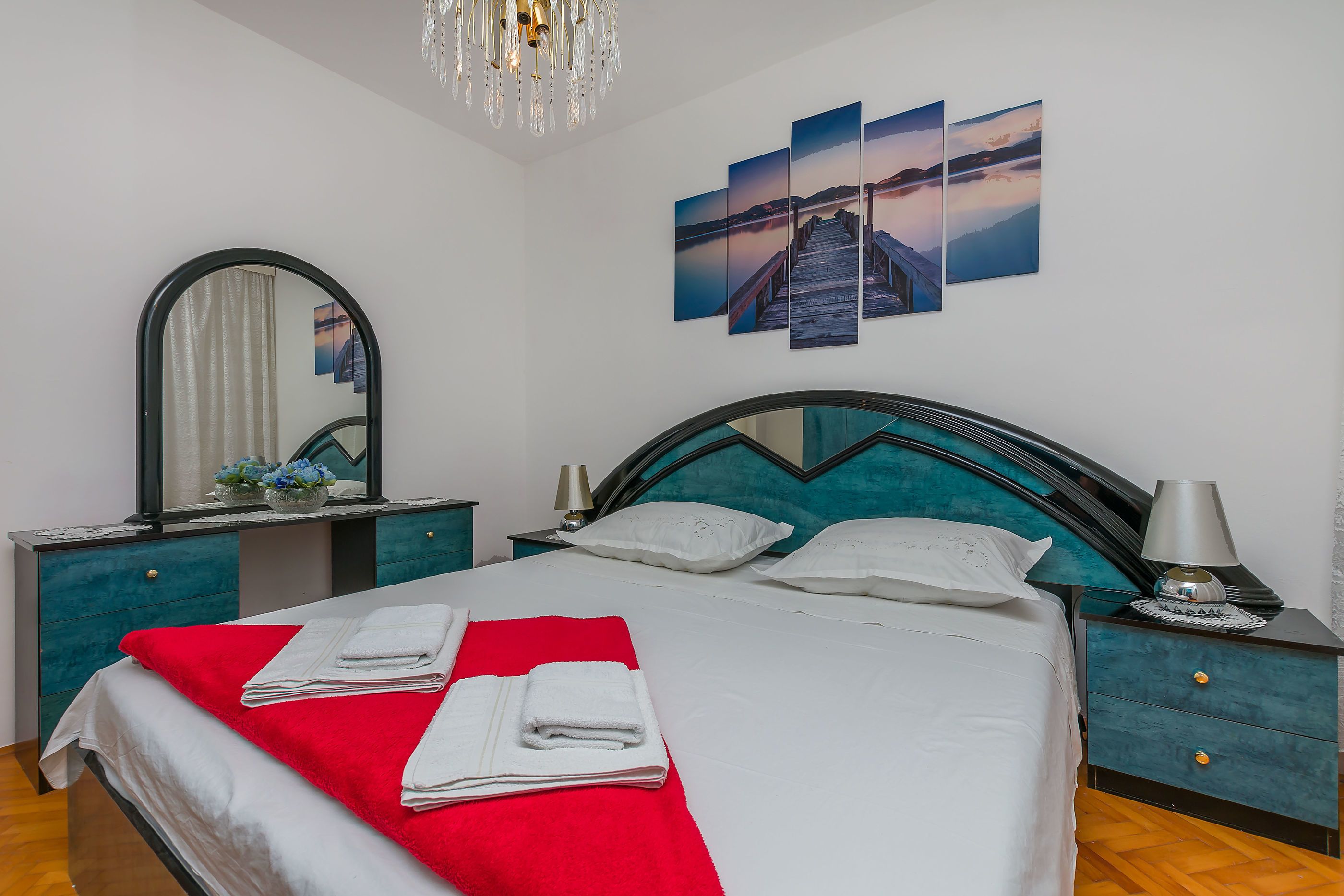 A neatly made bed with white linens and a red blanket, a large mirror, a nightstand with a lamp, and a wall with three sunset paintings.