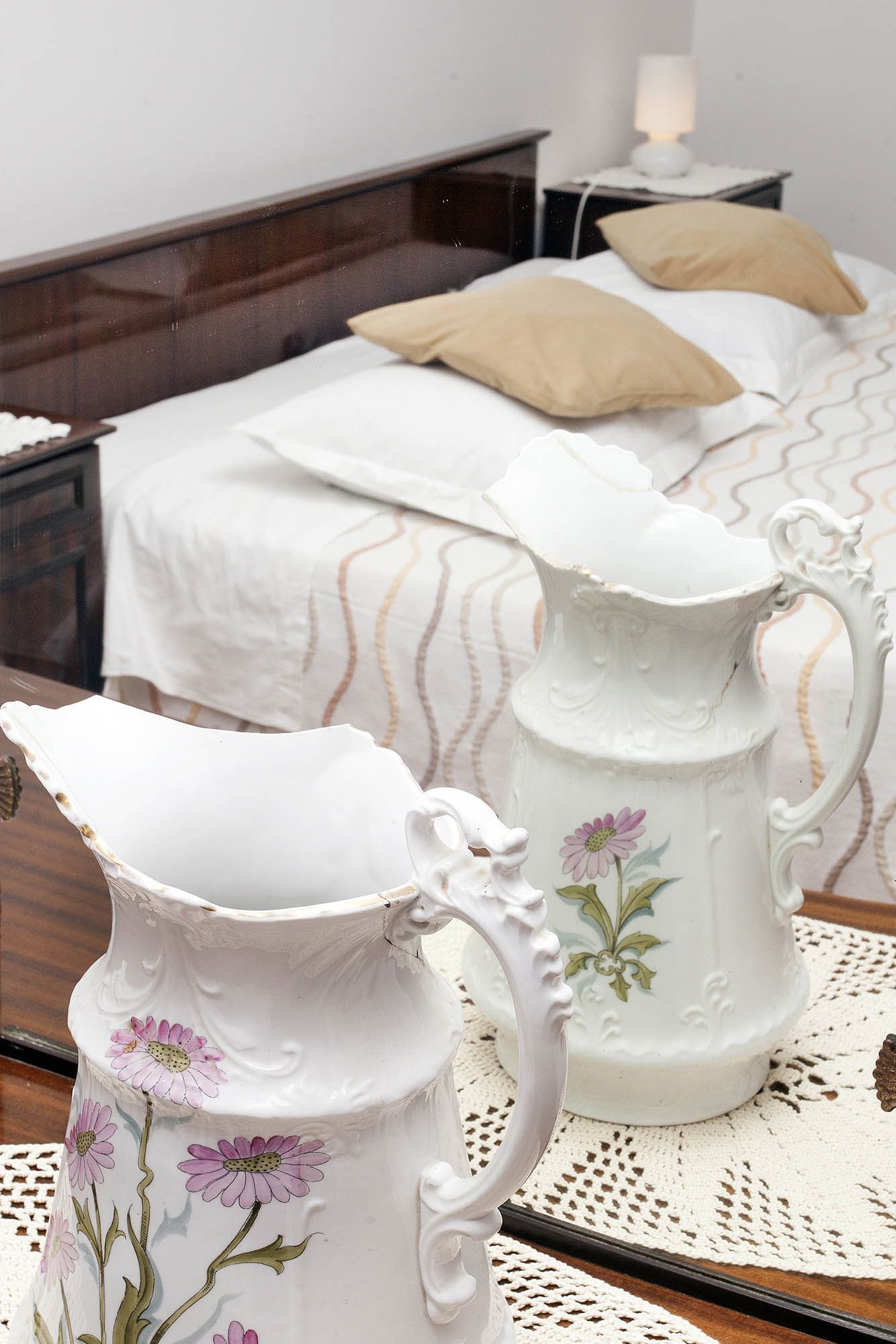 A bedroom with a bed, a nightstand, and two white ceramic pitchers with floral designs.