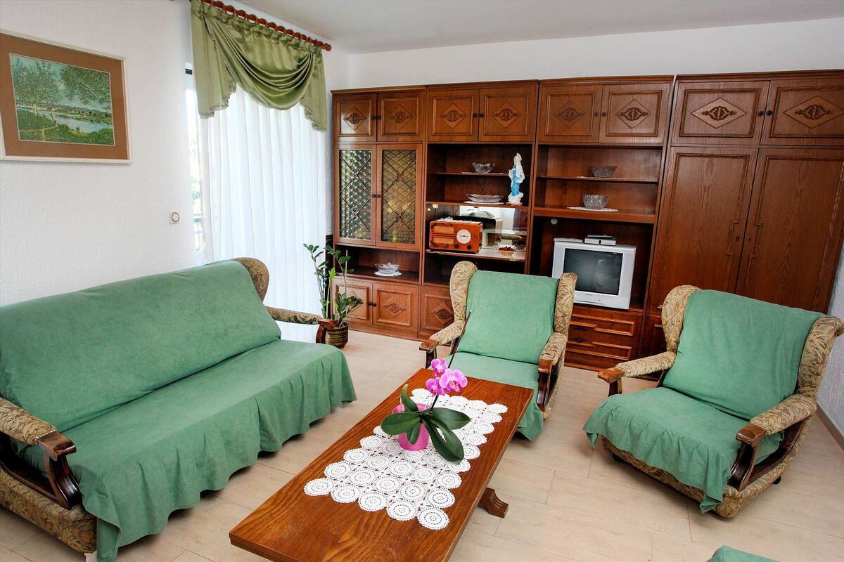 Un salon avec un canapé vert, une table basse en bois ornée d’un chemin de table en dentelle et un téléviseur posé sur un meuble en bois.