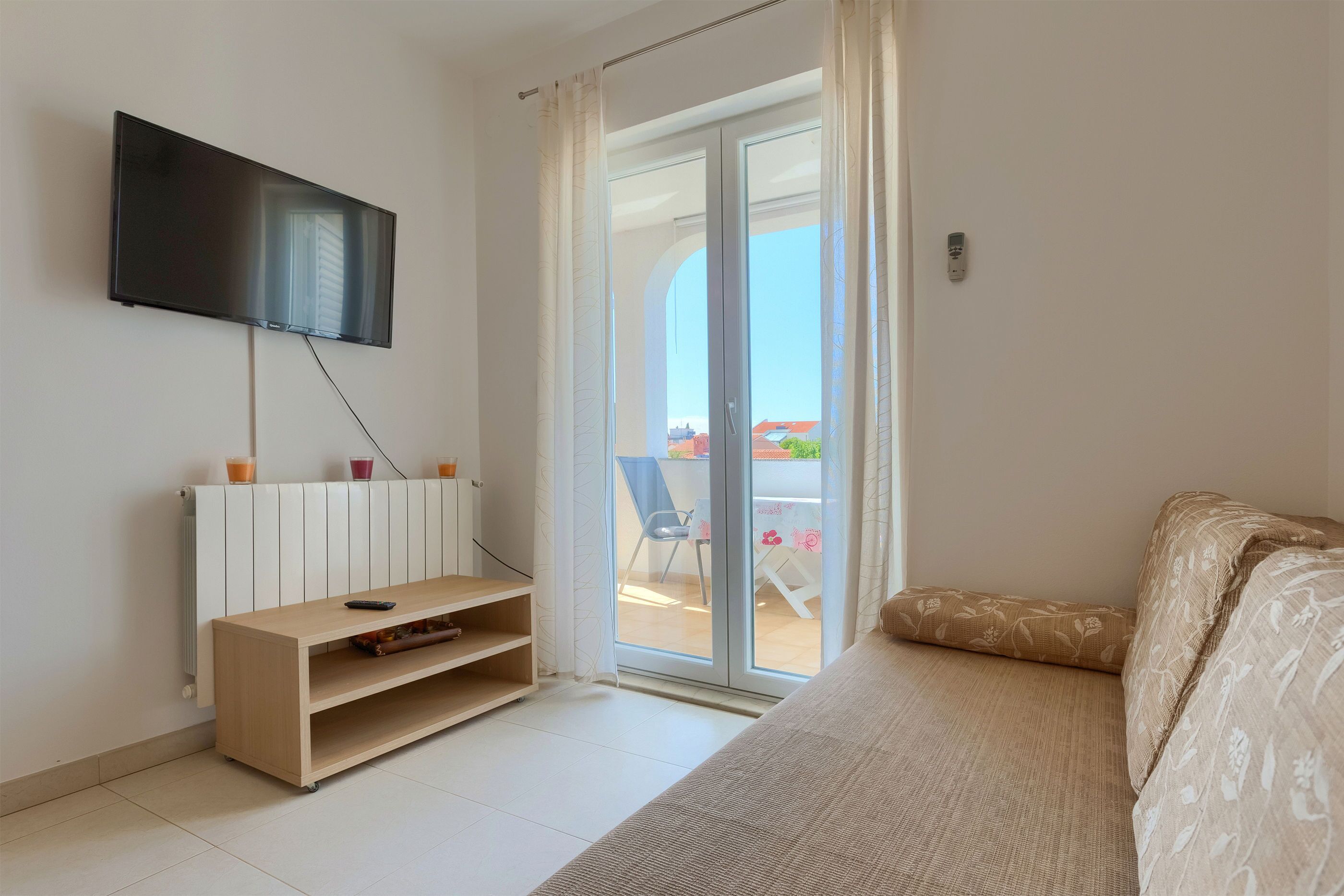 A modern room with a flat-screen TV mounted on the wall, a wooden TV stand, a sofa, and a sliding glass door leading to an outdoor area with a table and chairs.