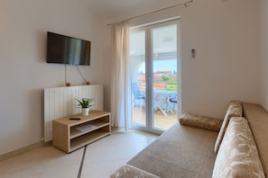 A modern living room with a sofa, a TV mounted on the wall, a small wooden table with a plant, and a sliding glass door leading to an outdoor area. - Apartments Boris (Fazana)